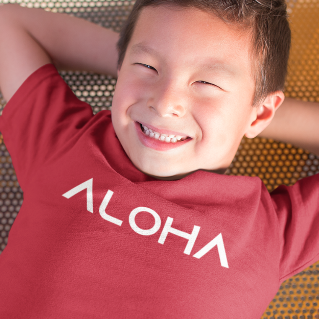 A young boy smiling while wearing a red minimalist Kids Aloha Shirt from Simply Aloha On The Go.