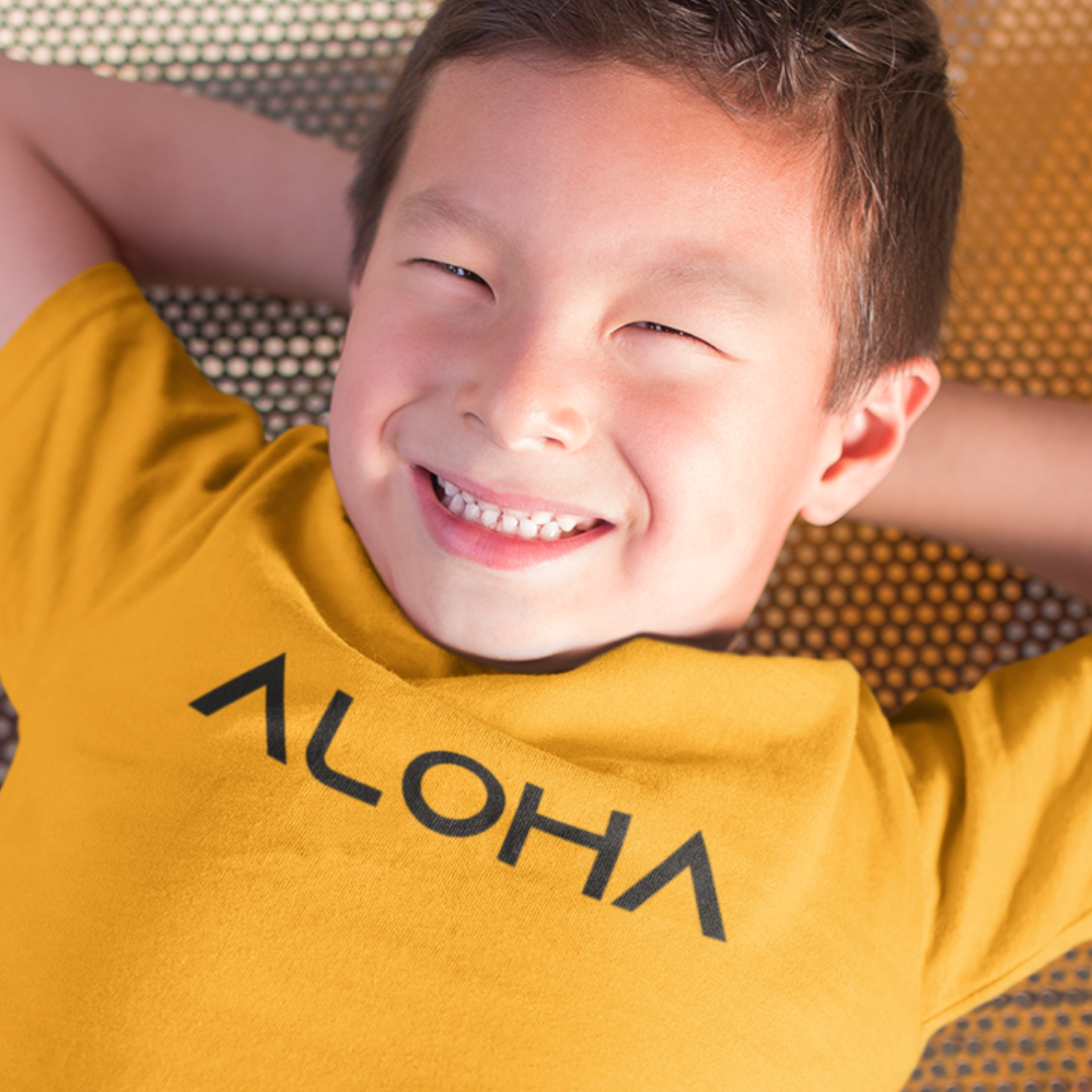A young boy smiling while wearing a yellow gold minimalist Kids Aloha Shirt from Simply Aloha On The Go.