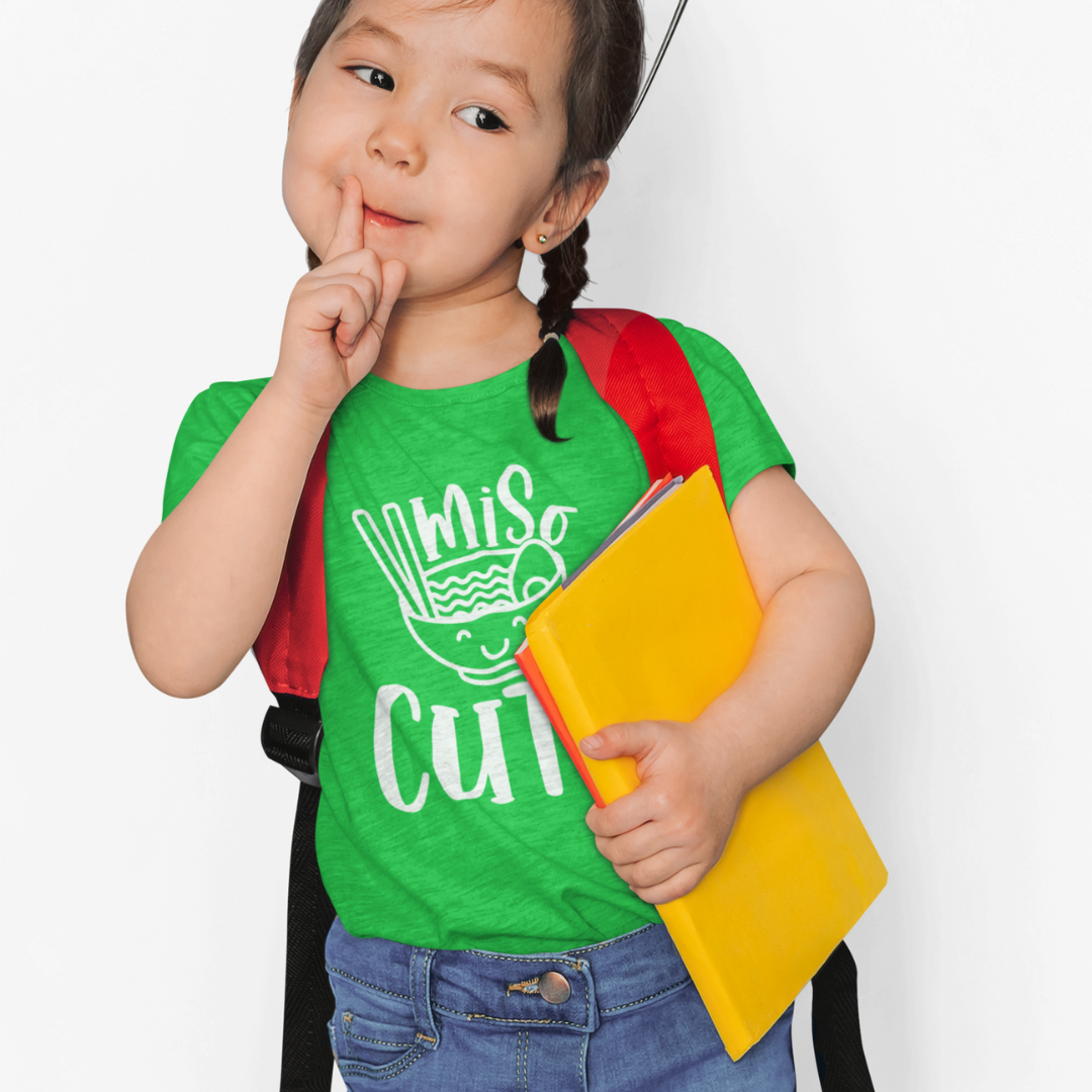 A young girl wearing a green t-shirt that says "Miso Cute" with a graphic of a smiling bowl of miso soup in a kawaii style. Available in different colors and sizes
