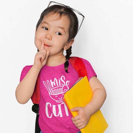 A young girl wearing a pink t-shirt that says "Miso Cute" with a graphic of a smiling bowl of miso soup in a kawaii style. Available in different colors and sizes
