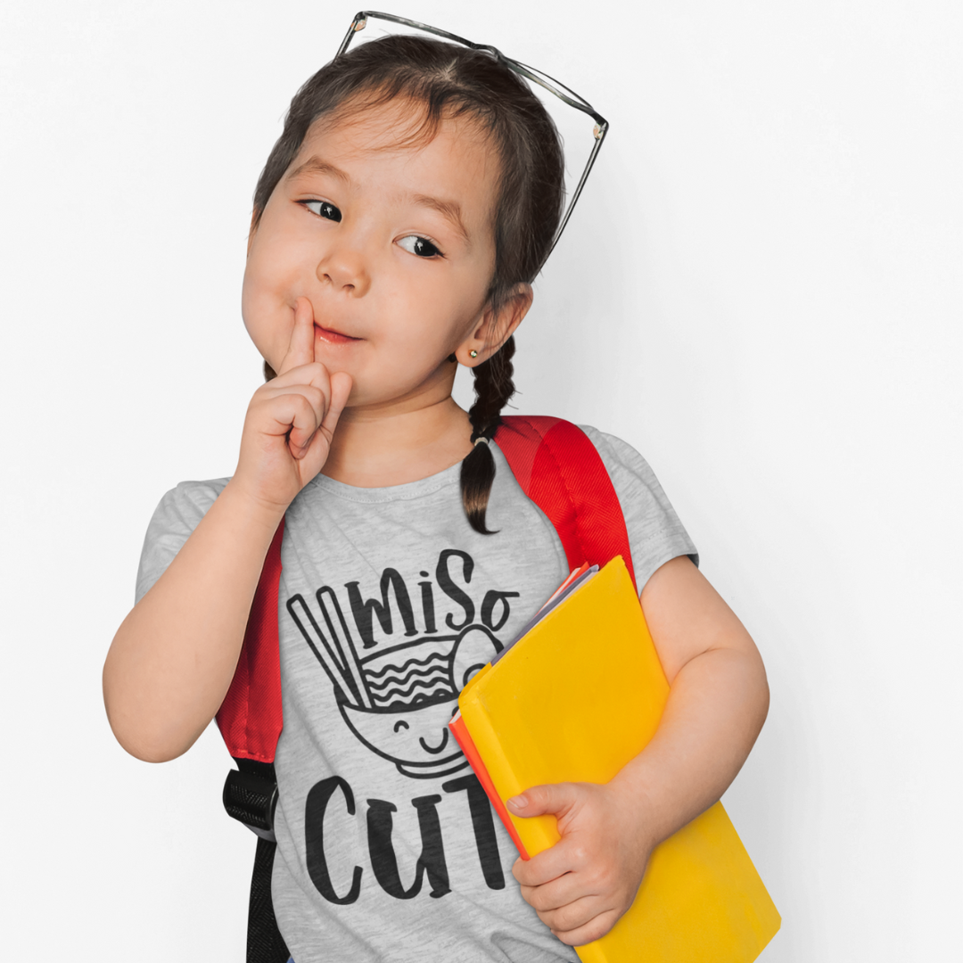 A young girl wearing a grey t-shirt that says "Miso Cute" with a graphic of a smiling bowl of miso soup in a kawaii style. Available in different sizes and colors
