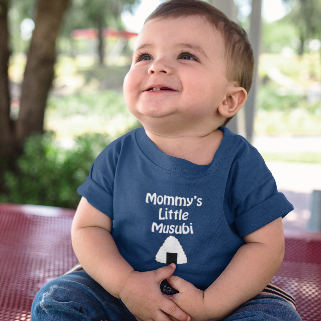Child wearing a blue shirt with "Mommy's Little Musubi" text, sitting outdoors.