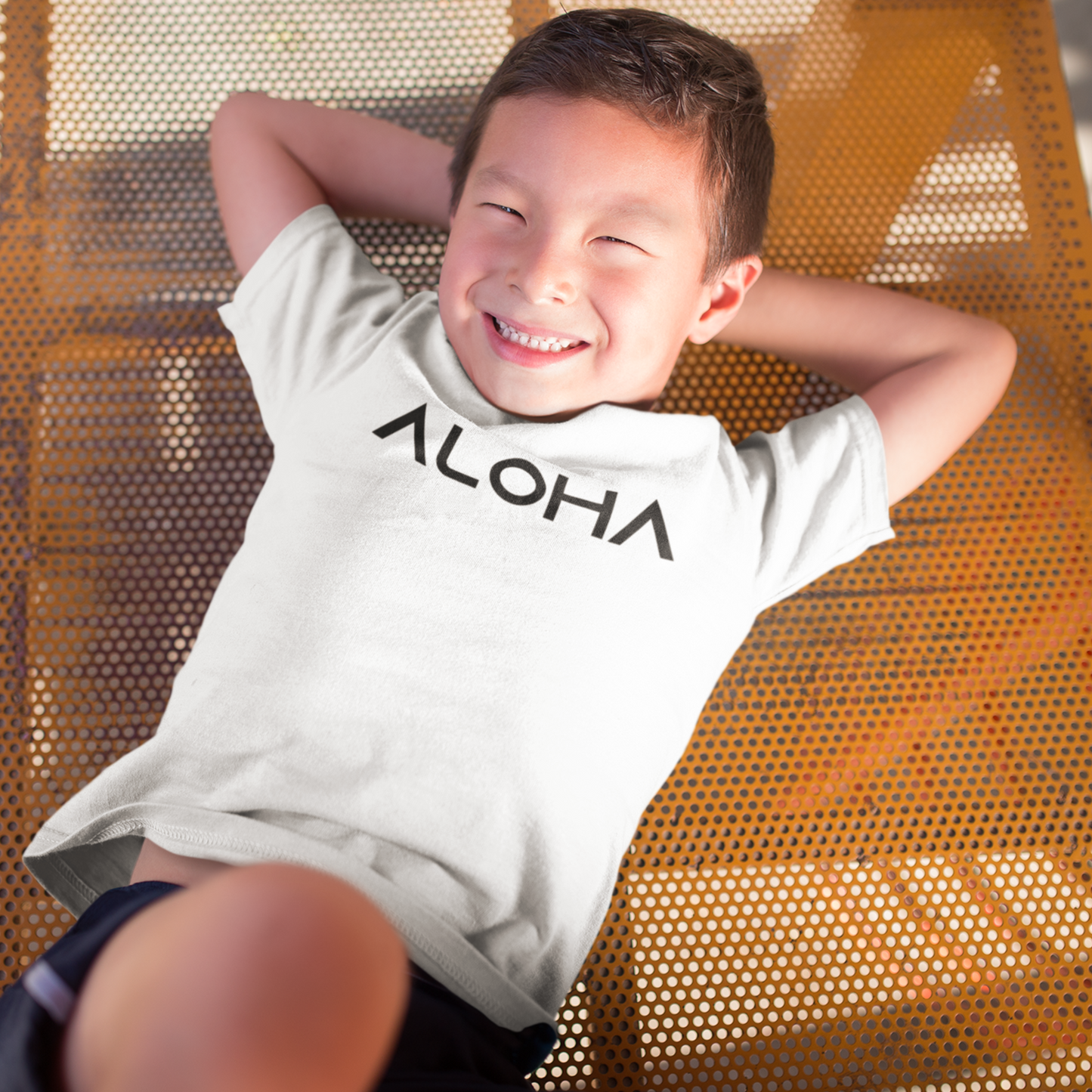 A young child wearing a white t-shirt that says "Aloha" in a black font, a Hawaiian-inspired shirt for kids. Available in different tropical colors and sizes