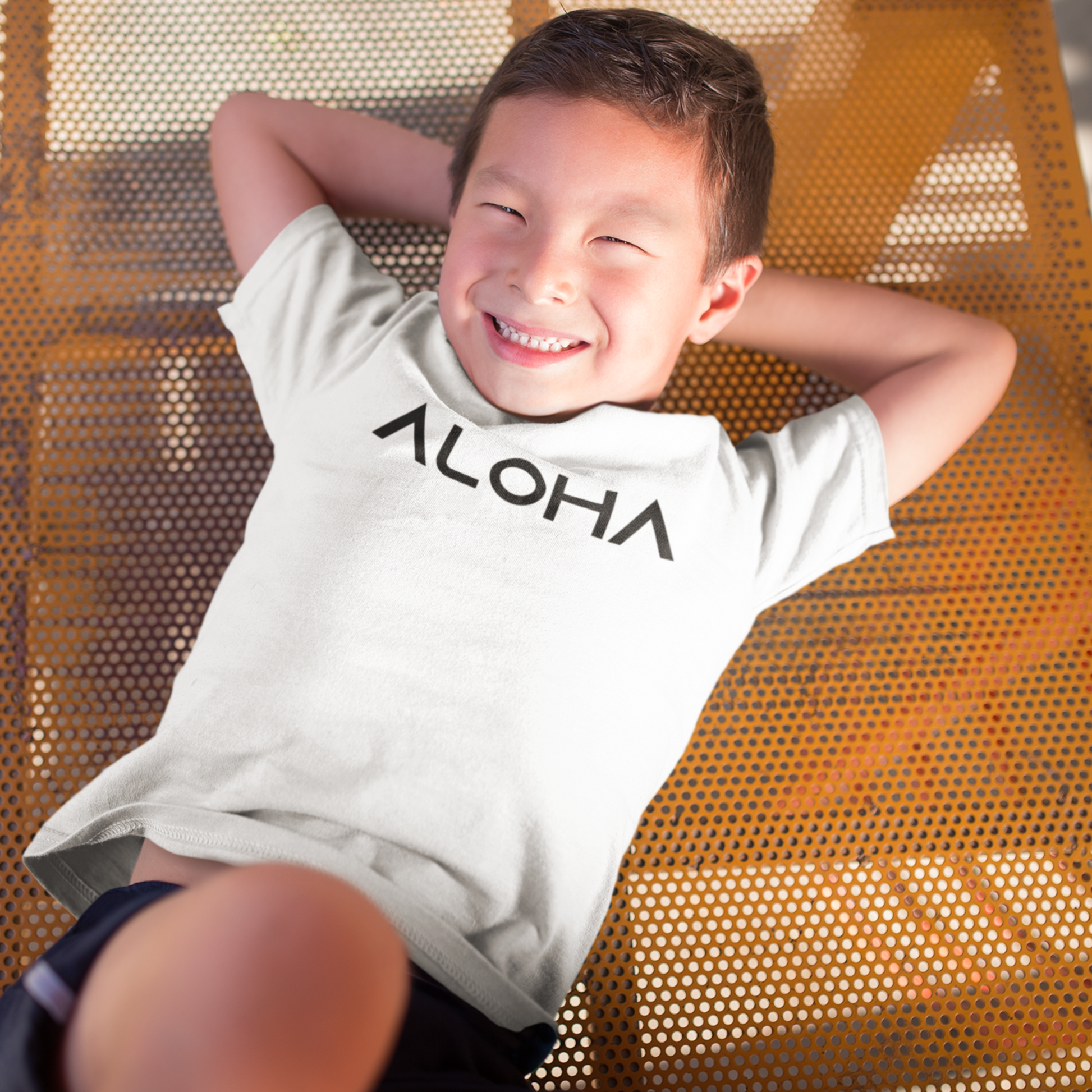 A young child wearing a white t-shirt that says "Aloha" in a black font, a Hawaiian-inspired shirt for kids. Available in different tropical colors and sizes