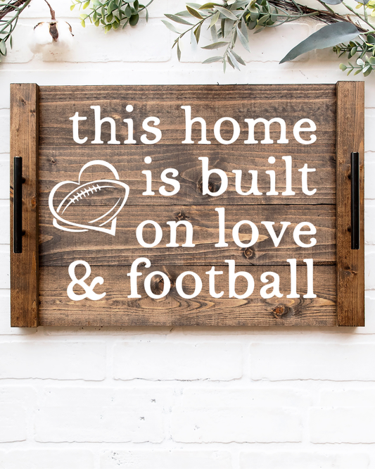 Rustic dark walnut wood noodle board stove cover with white painted text reading this This Home Is Built On Love & Football with a heart graphic and black metal handles