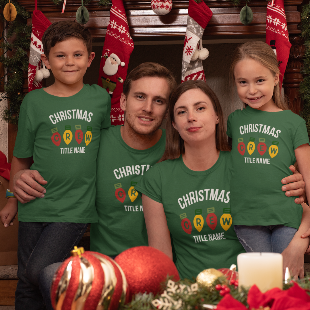 A smiling family of four sitting by a Christmas tree wearing matching custom white holiday t-shirts with festive light bulb graphics.