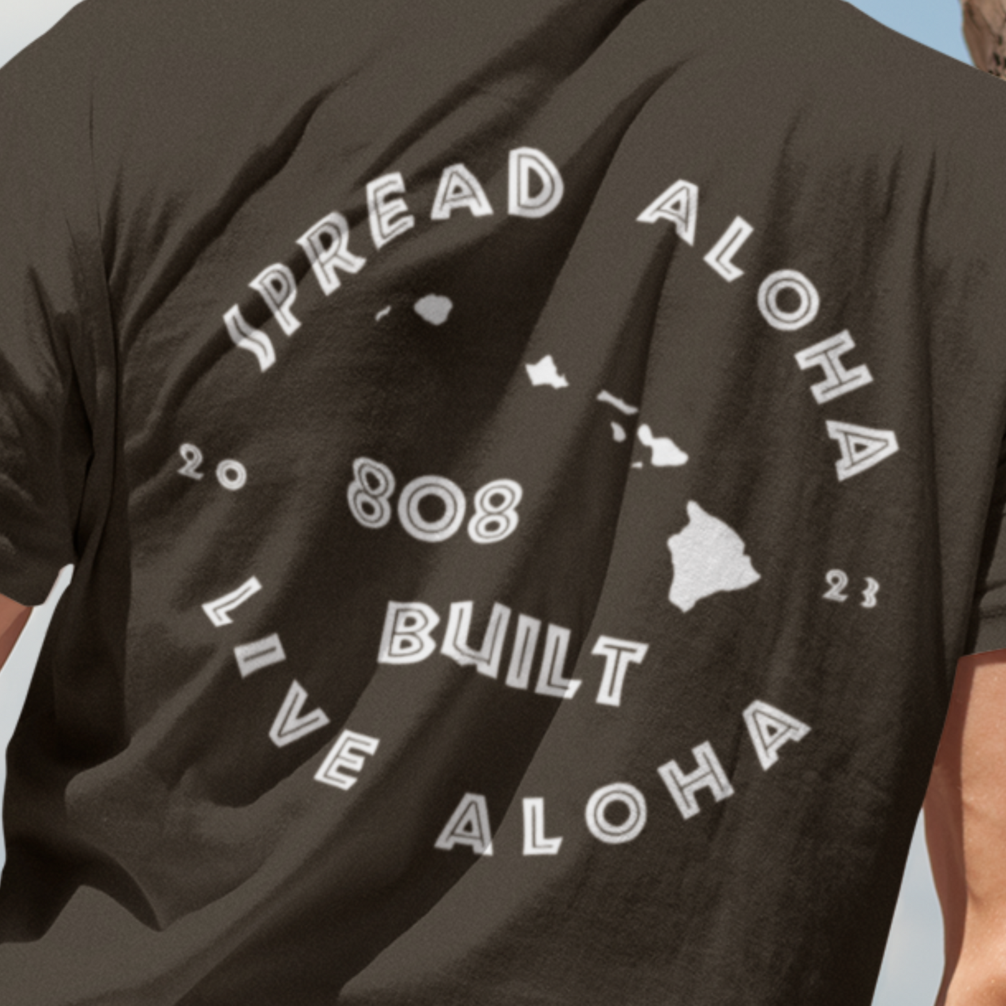 Back view of a man on a beach wearing a brown 808 Built Hawaii t-shirt with a white circular graphic that reads Spread Aloha Live Aloha 808 Built
