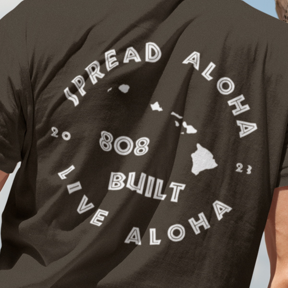 Back view of a man on a beach wearing a brown 808 Built t-shirt with a white circular graphic that reads Spread Aloha Live Aloha 808 Built