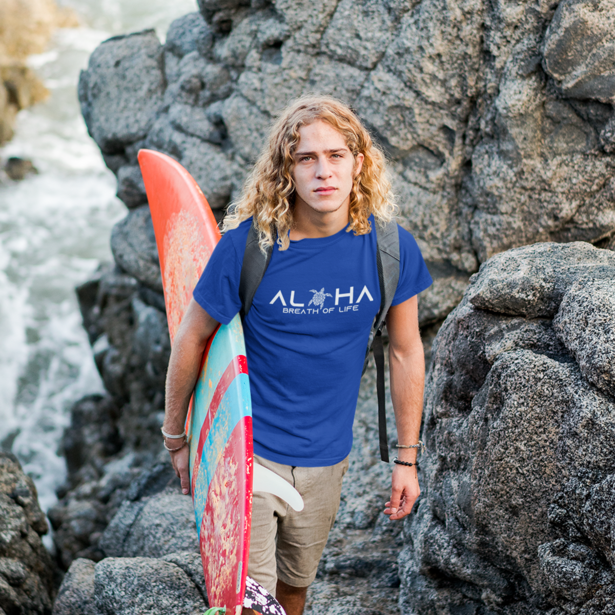 Man wearing a blue Men's Breath of Life T-Shirt, Hawaiian-inspired tee featuring a honu sea turtle while holding a colorful surfboard.