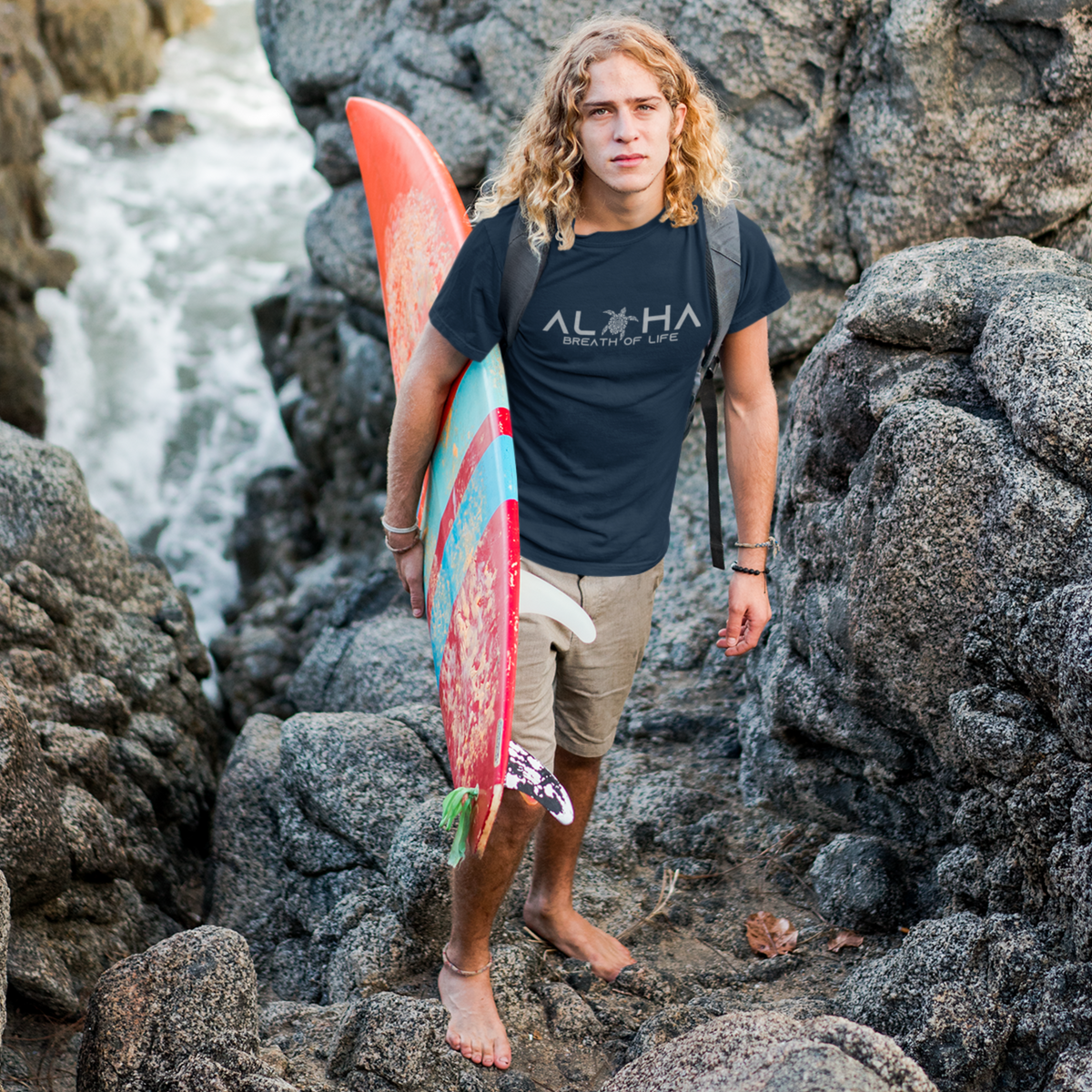 Man wearing anavy blue Men's Breath of Life T-Shirt, Hawaiian-inspired tee featuring a honu sea turtle while holding a colorful surfboard.