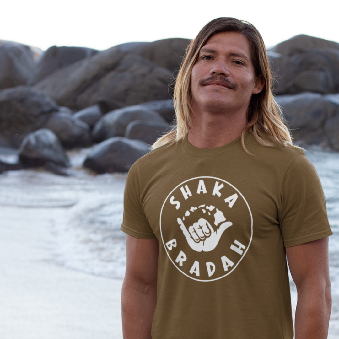 A man outdoors wearing a custom Shaka Bradah T-Shirt in brown, white font with a graphic of a hand making the shaka sign.