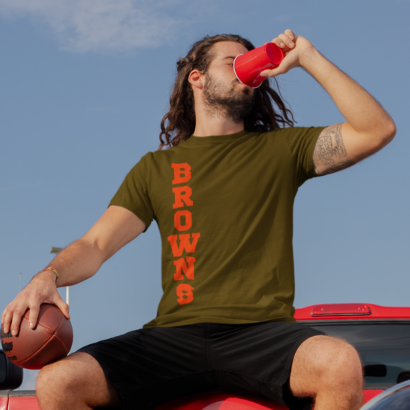 A man at a tailgate party wearing a personalized brown men's game day t-shirt featuring vertical blue text reading "BROWNS"