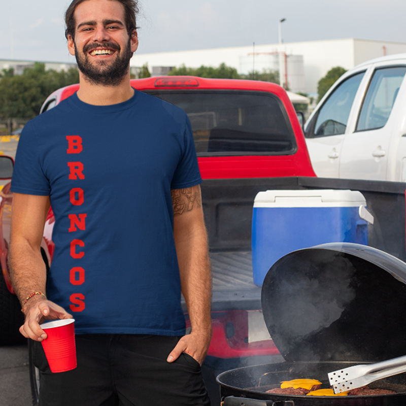 A man at a tailgate party wearing a personalized men's game day t-shirt featuring vertical orange text reading "BRONCOS"