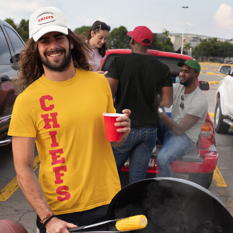 A man at a tailgate party wearing a personalized yellow men's game day t-shirt featuring vertical red text reading "CHIEFS"