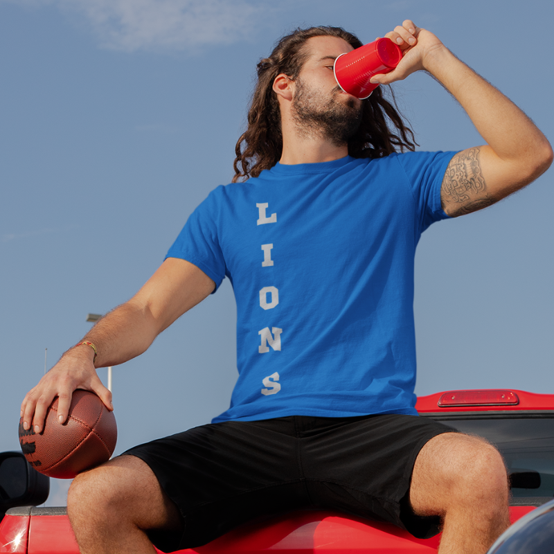 A man at a tailgate party wearing a personalized blue men's game day t-shirt featuring vertical blue text reading "LIONS"