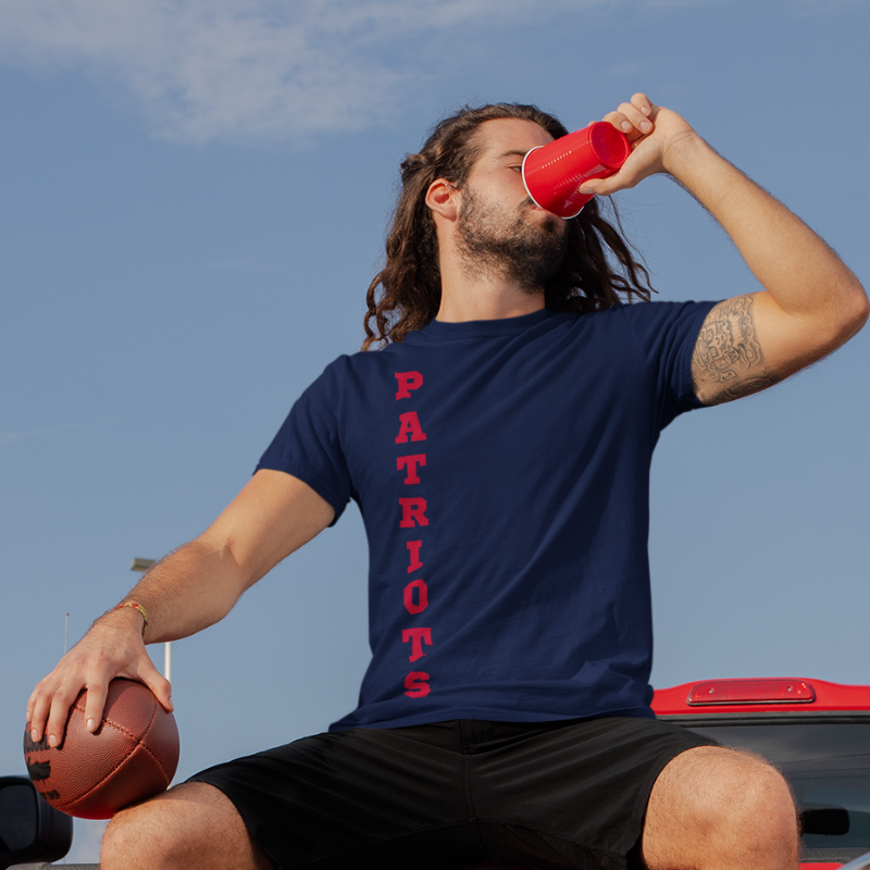 A man at a tailgate party wearing a personalized nautical blue men's game day t-shirt featuring vertical red text reading "PATRIOTS"
