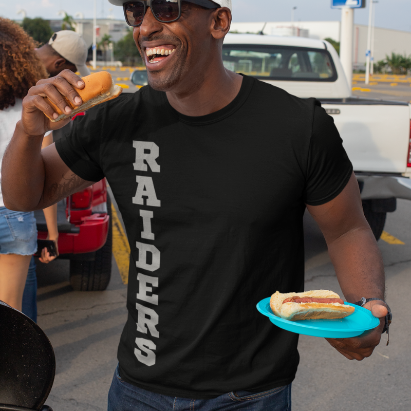 A man at a tailgate party wearing a personalized black men's game day t-shirt featuring vertical silver text reading "RAIDERS"