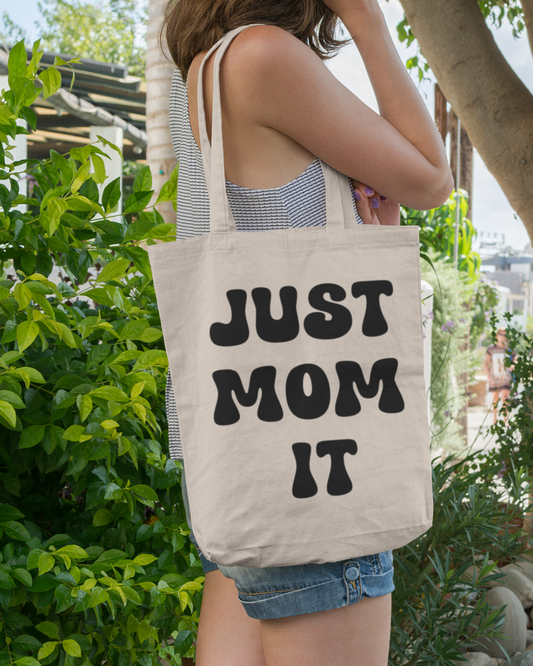 Person holding a tote bag with "JUST MOM IT" text in an outdoor setting