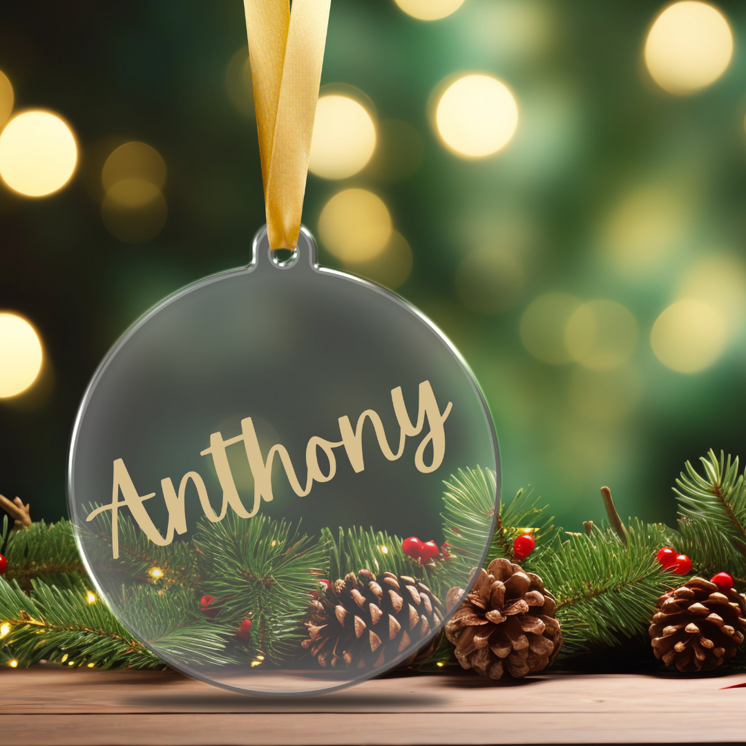 A clear, flat acrylic Christmas ornament personalized with a name in gold script font, hanging from a tree branch designed by Simply Aloha On The Go.