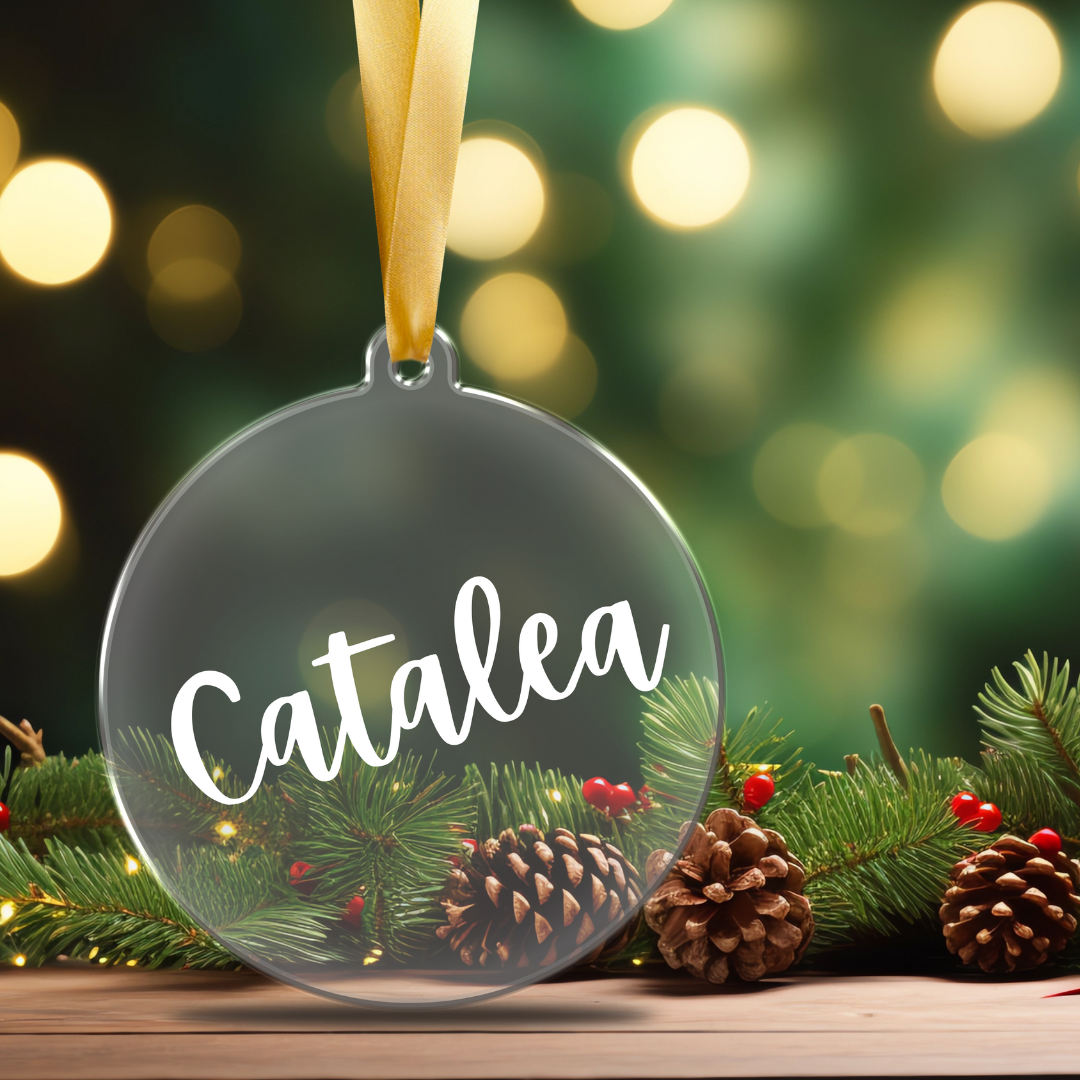 A clear, flat acrylic Christmas ornament personalized with a name  in white script font, hanging from a tree branch designed by Simply Aloha On The Go.