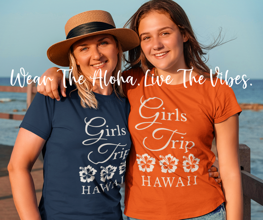 Two women smiling outdoors by the water wearing matching custom girls trip t-shirts in navy blue and bright orange that read Wear The Aloha Live The Vibes.