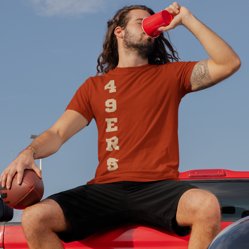 A man at a tailgate party wearing a personalized men's game day t-shirt featuring vertical gold text reading "49ERS"