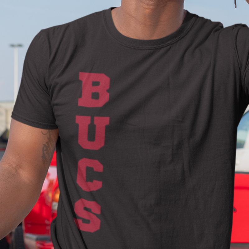 A man at a tailgate party wearing a personalized black men's game day t-shirt featuring vertical red text reading "BUCS"