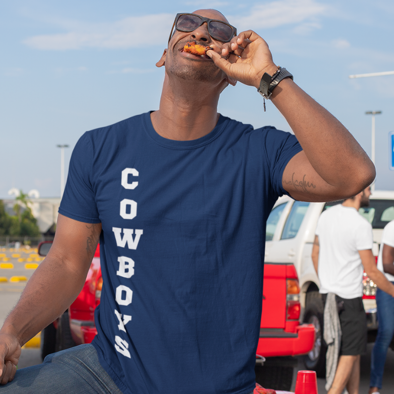 A man at a tailgate party wearing a personalized blue men's game day t-shirt featuring vertical white text reading "COWBOYS"