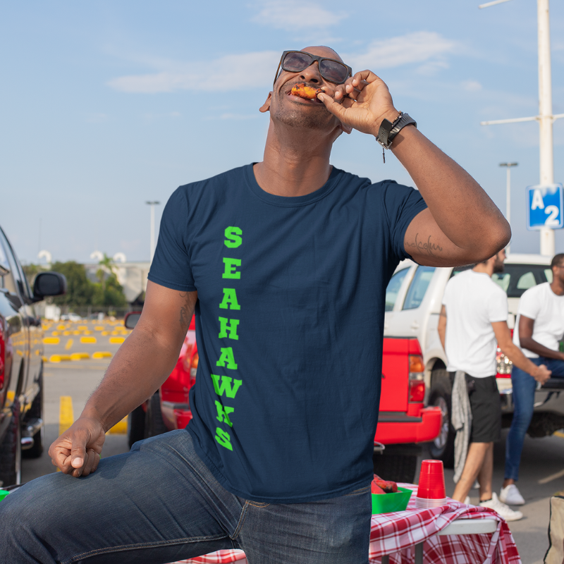 A man at a tailgate party wearing a personalized men's game day t-shirt featuring vertical blue text reading "SEAHAWKS"