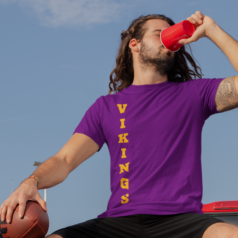 A man at a tailgate party wearing a personalized men's game day t-shirt featuring vertical gold text reading "VIKINGS"