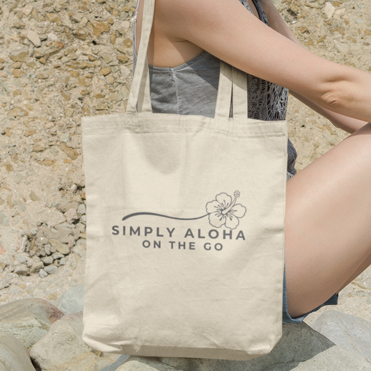 Reusable canvas tote bag featuring the double-sided Simply Aloha On The Go hibiscus logo in gray, carried over the shoulder in a natural outdoor setting.