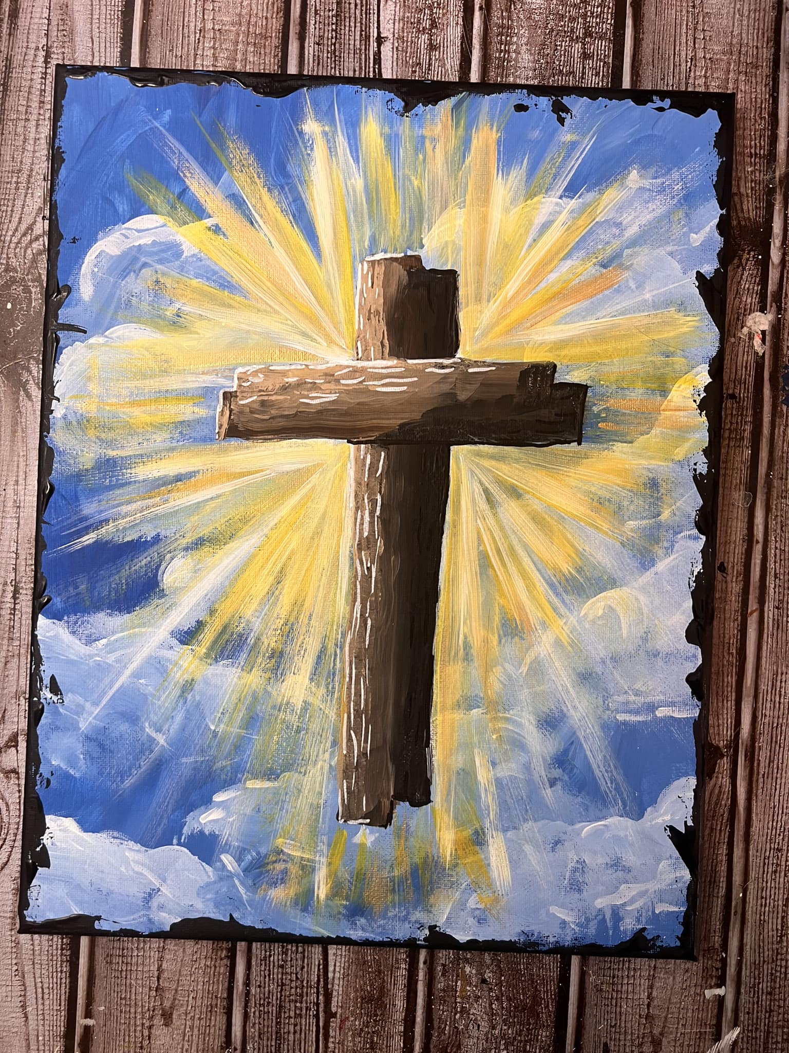 A completed painting from the rugged cross DIY Easter kit, featuring a brown wooden cross against a bright blue and white cloudy sky with radiating yellow light beams.
