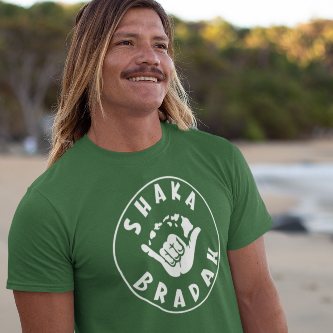 A man wearing a green men's t-shirt that says "Shaka Bradah" in a simple white font with a graphic of a hand making the shaka sign.
