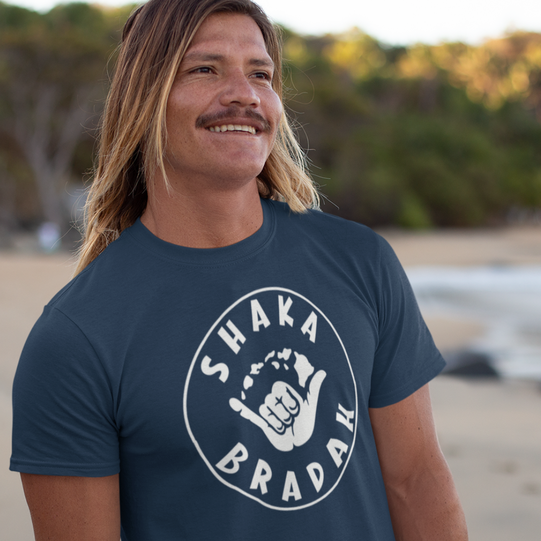 A man wearing a blue men's t-shirt that says "Shaka Bradah" in a simple white font with a graphic of a hand making the shaka sign.