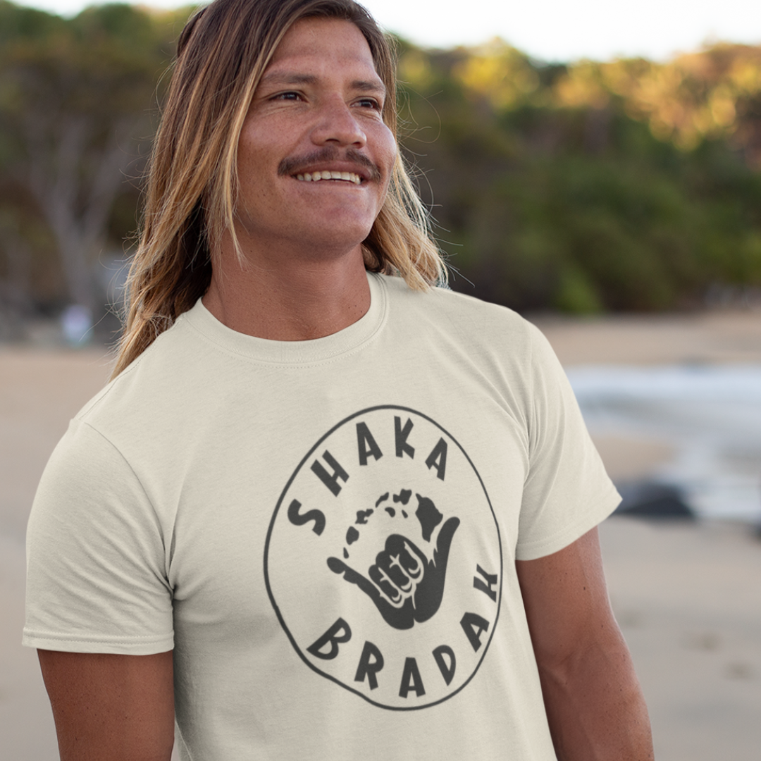 A man wearing a sand men's t-shirt that says "Shaka Bradah" in a simple white font with a graphic of a hand making the shaka sign.