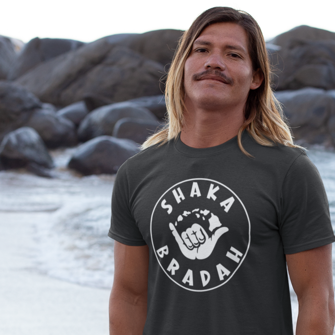 A man wearing a black men's t-shirt that says "Shaka Bradah" in a simple white font with a graphic of a hand making the shaka sign.