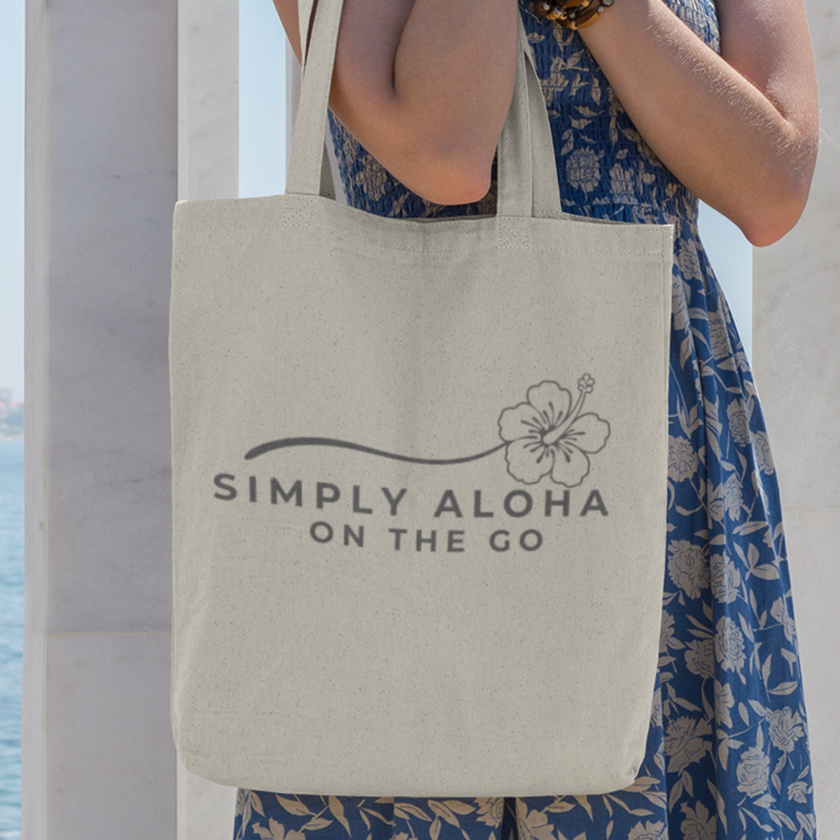Reusable canvas tote bag featuring the double-sided Simply Aloha On The Go hibiscus logo in gray, carried over the shoulder in a natural outdoor setting.