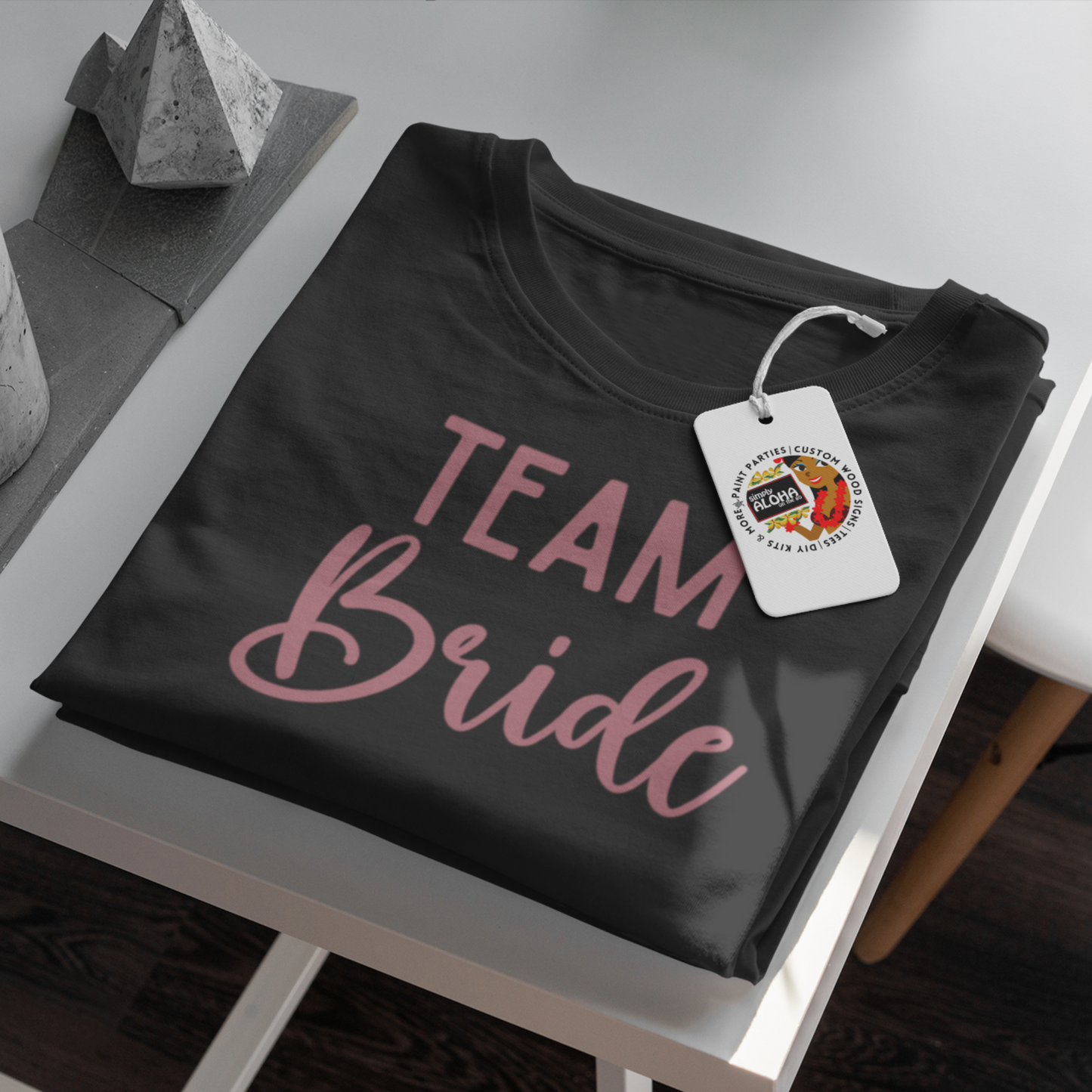 A closeup look of a clean and chic black t-shirt with the word "Team Bride" printed in a subtle, elegant pink font. The shirt is a must-have for a bridal shower or bachelorette party