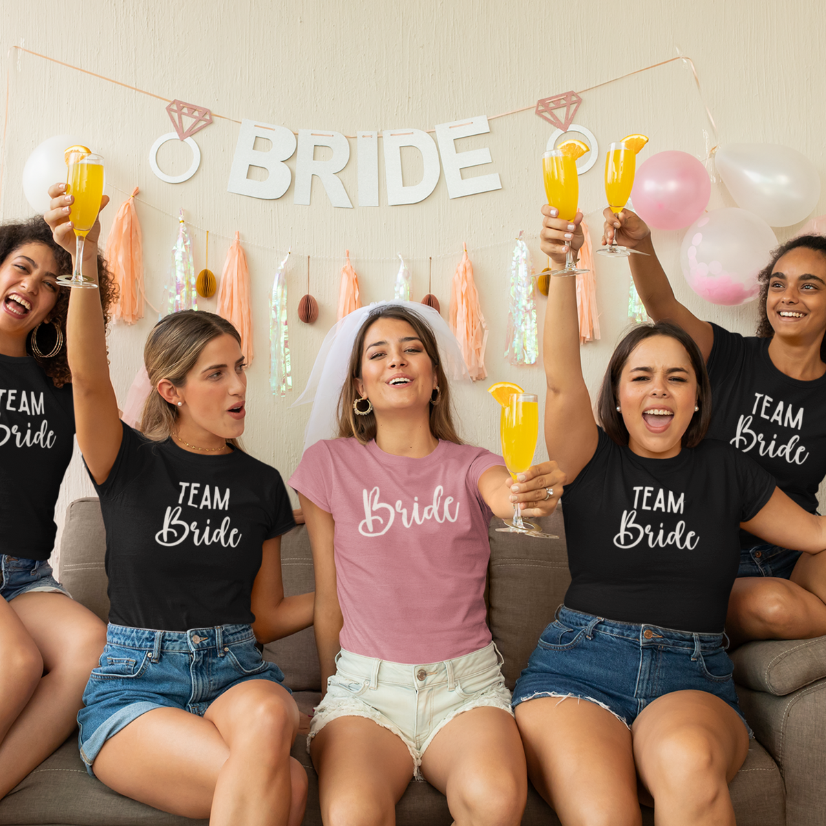 5 women on their Bride Tribe Shirt celebrating at an indoor bridal shower wearing matching pink Bride and black Team Bride bachelorette party shirts.