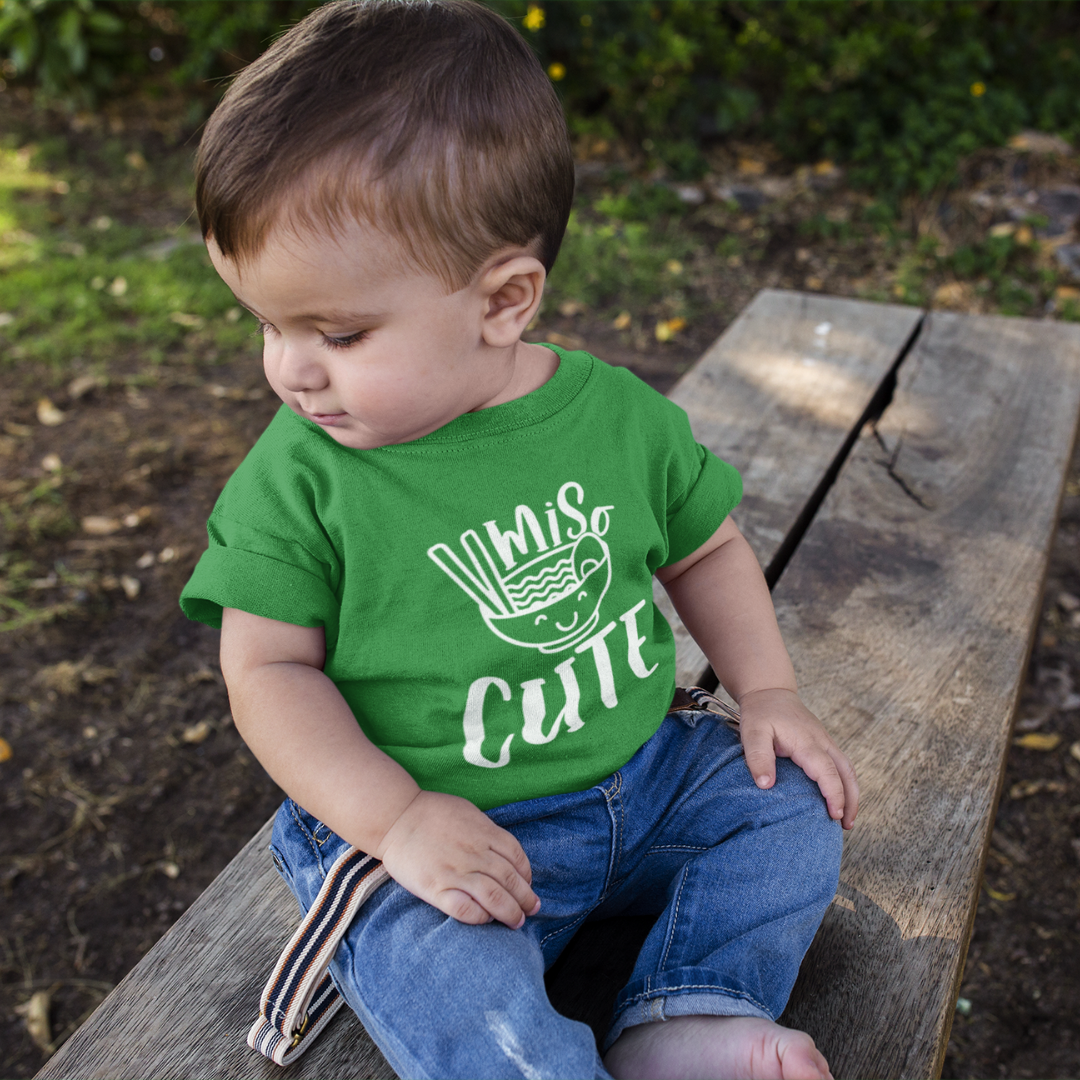 A young boy sitting outside on a stone wall wearing a green custom kids t-shirt with a white graphic that reads Miso Cute.