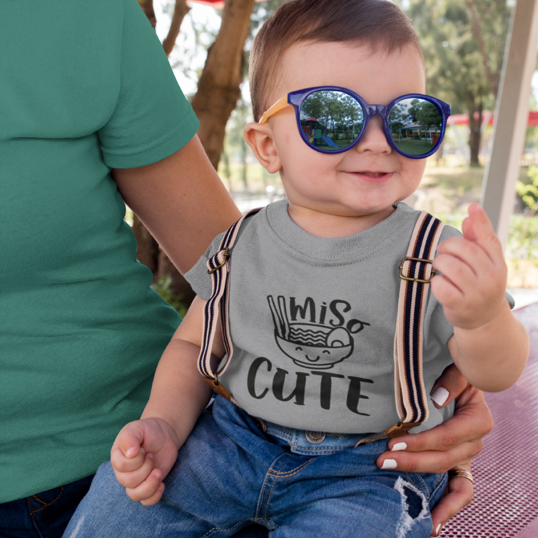 Baby wearing sunglasses and a t-shirt with Miso Cute text, and graphic of a bowl with chopsticks held by an adult outdoors.