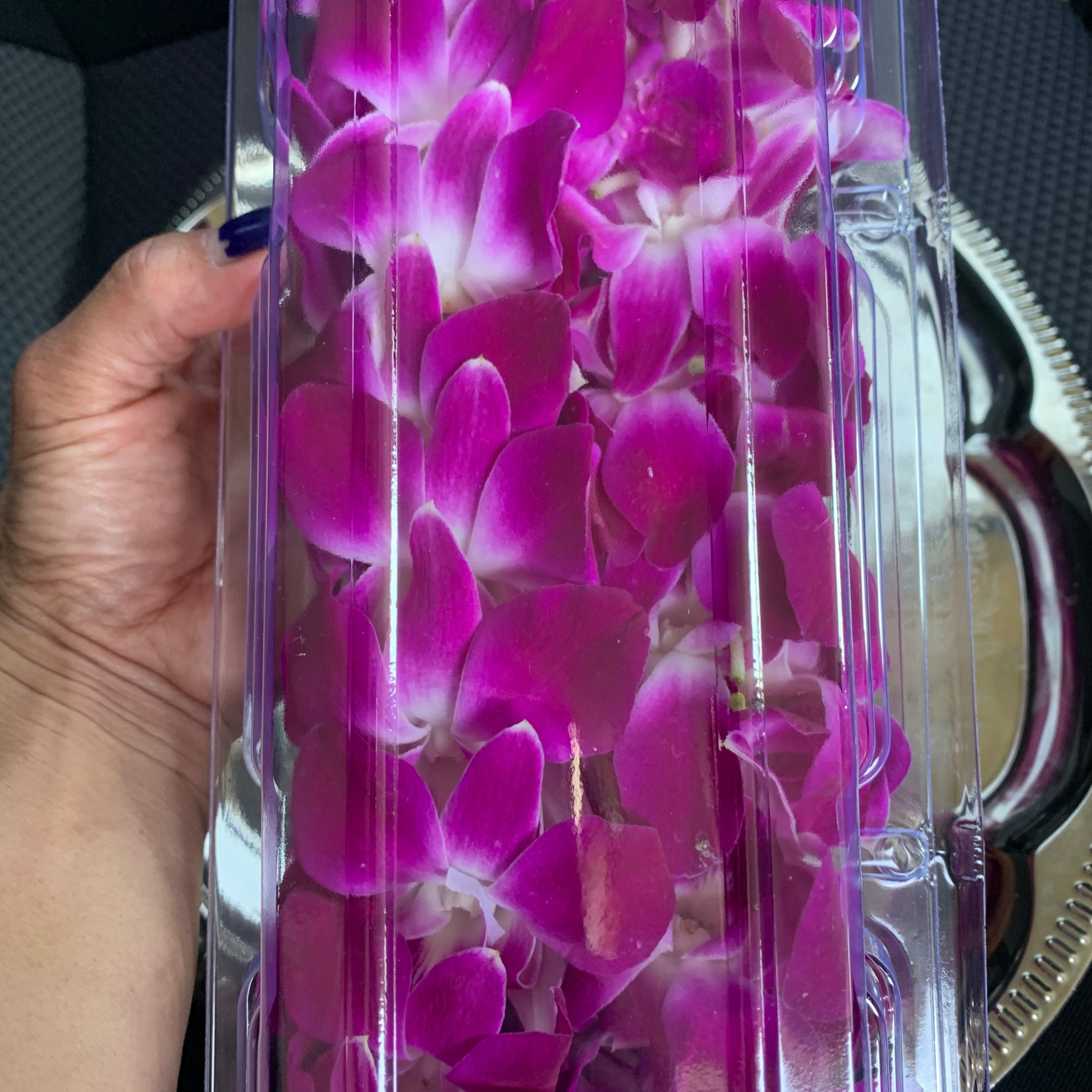 A close-up shot of a person's hands holding a single fresh purple orchid lei. The lei is individually packaged delicate blossoms -hand-strung in Arizona.