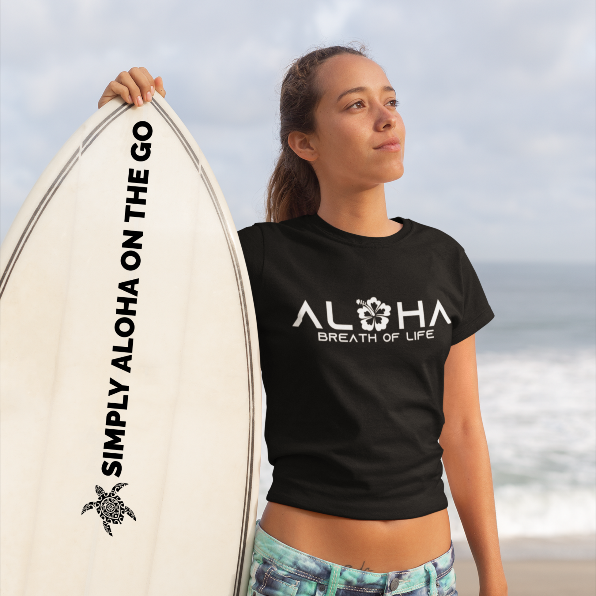 A woman on a beach holding a surfboard branded with Simply Aloha On The Go while wearing a black Aloha Breath of Life t-shirt.
