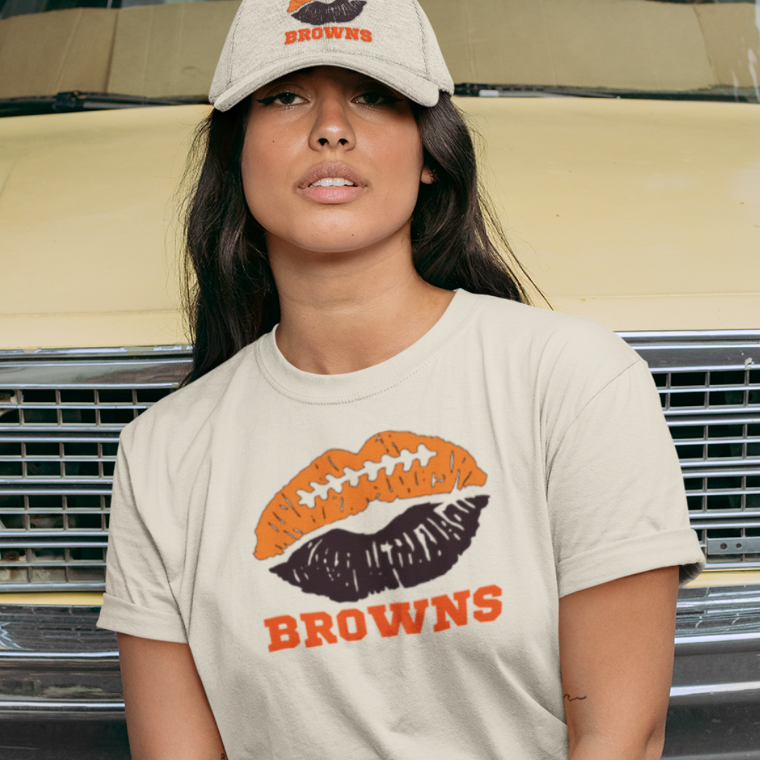A woman wearing football game day t-shirt in cream color with a lip-themed design in brown and ornageon top and the team name Browns written below.