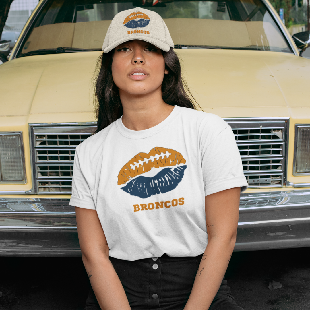 A woman wearing football game day t-shirt in cream color with a lip-themed design in navy blue and orange on top and the team name Broncos written below.
